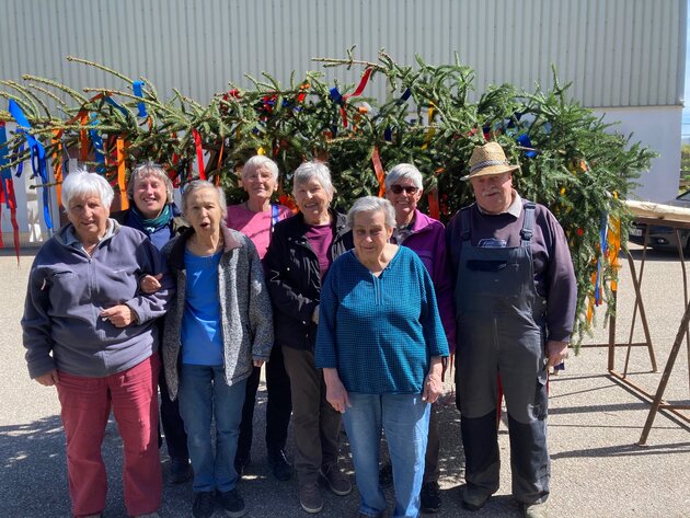 Eine Gruppe Frauen steht vor dem liegenden Maibaum. Er ist mit bunten Bändern behangen. Rechts steht ein Mann mit Latzhose und Strohhut.