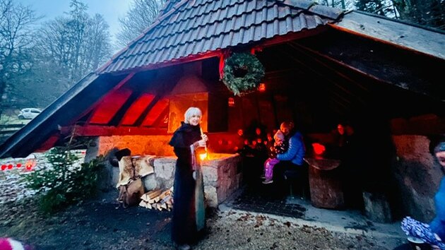 Vor der weihnachtlich dekorierten Grillhütte im Wald steht eine Märchenerzählerin und spielt Flöte. Um die Hütte herum ist ein stimmungsvolles Licht. 