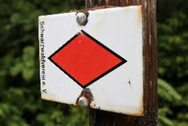 Das Bild zeigt ein altes Emailschild, das auf einem Holzpfosten angebracht ist. Es zeigt eine quer liegende rote Raute auf weißem Hintergrund.