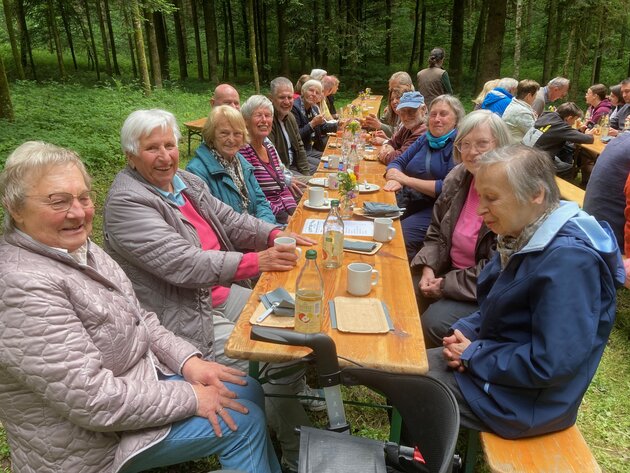 Eine Gruppe mit fröhlich schauenden Menschen sitzt auf einer Bierbank mitten im Wald. Vor ihnen stehen Getränke und Essen. 