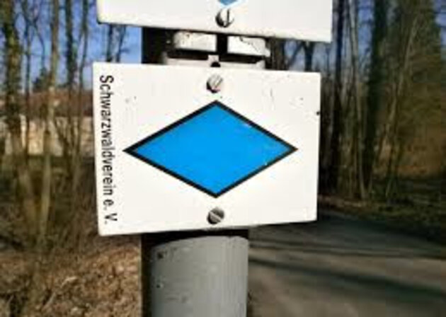 Das Bild zeigt ein weißes Schild mit quer liegender blauer Raute. Es ist an einen Metallpfosten montiert. Seitlich steht Schwarzwaldverein e.V. 